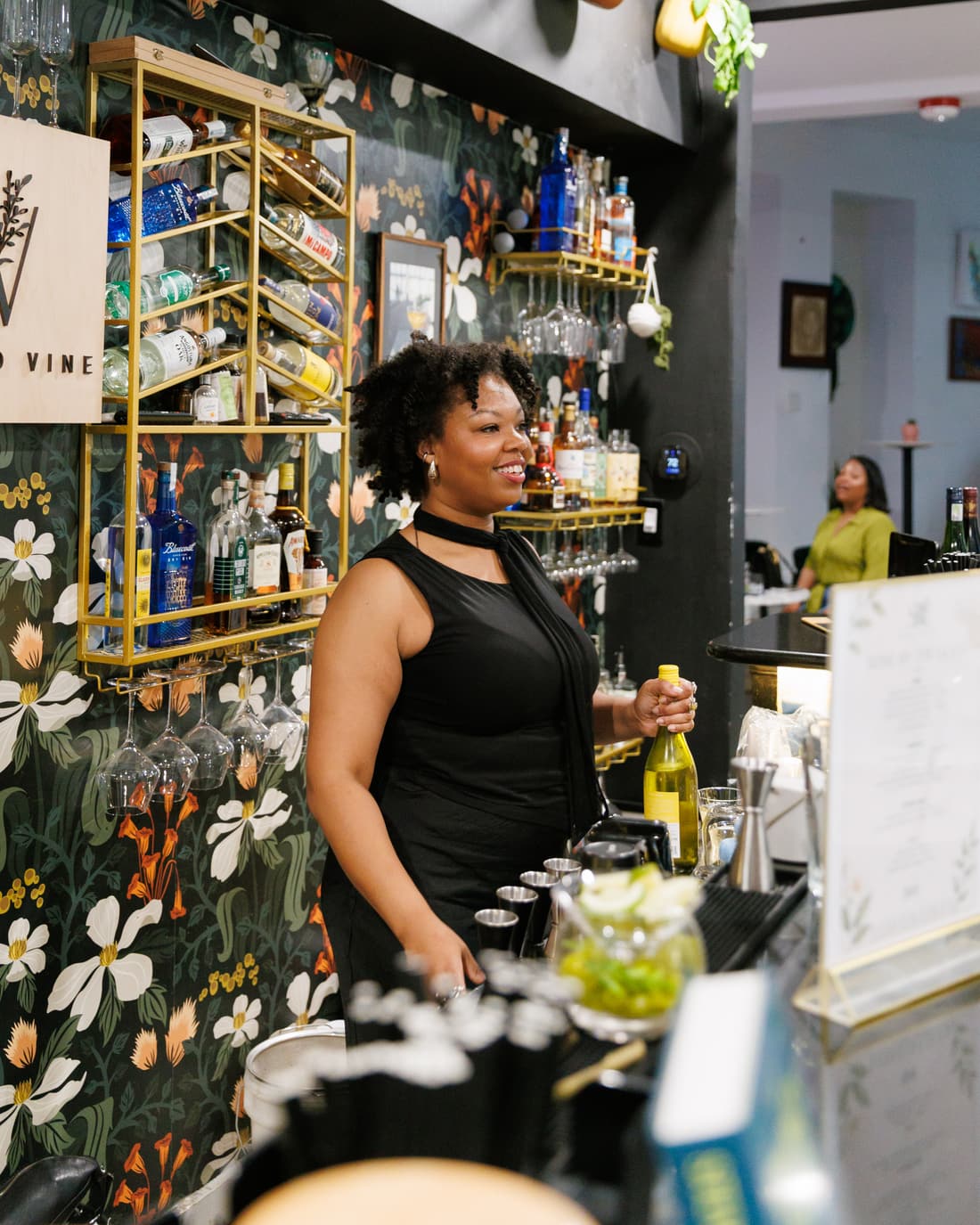 Bartender smiling behind the floral wallpaper and hanging glassware of the Botany Bar at Stem & Vine, Baltimore
