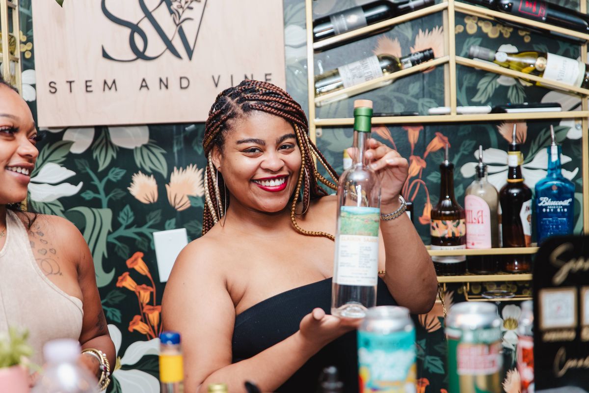 Bartender holding up a bottle beside the Stem & Vine sign at the floral wall bar in Baltimore