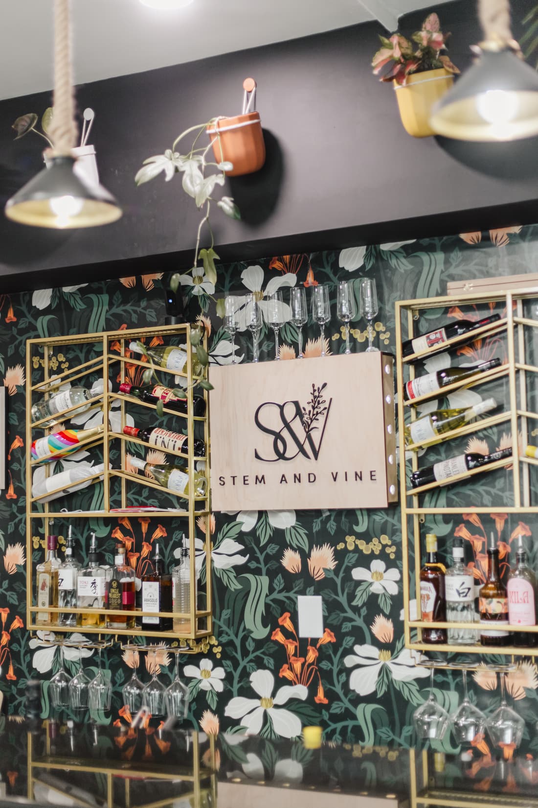 Brass shelves of wine and spirits framing the Stem & Vine sign against floral wallpaper in Baltimore