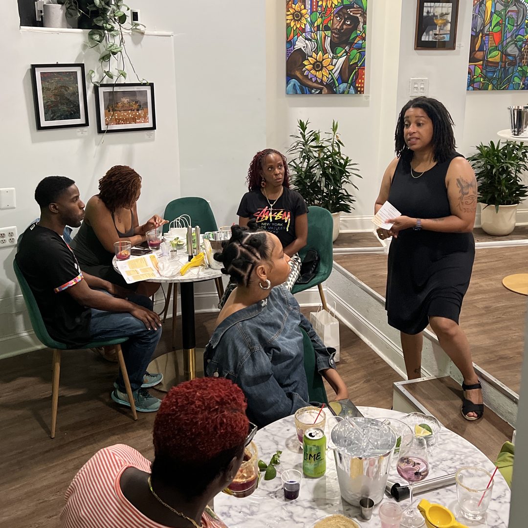 Brittany Williams talking a group through their drinks in the Gallery at Stem & Vine in Baltimore, original art on the walls behind them