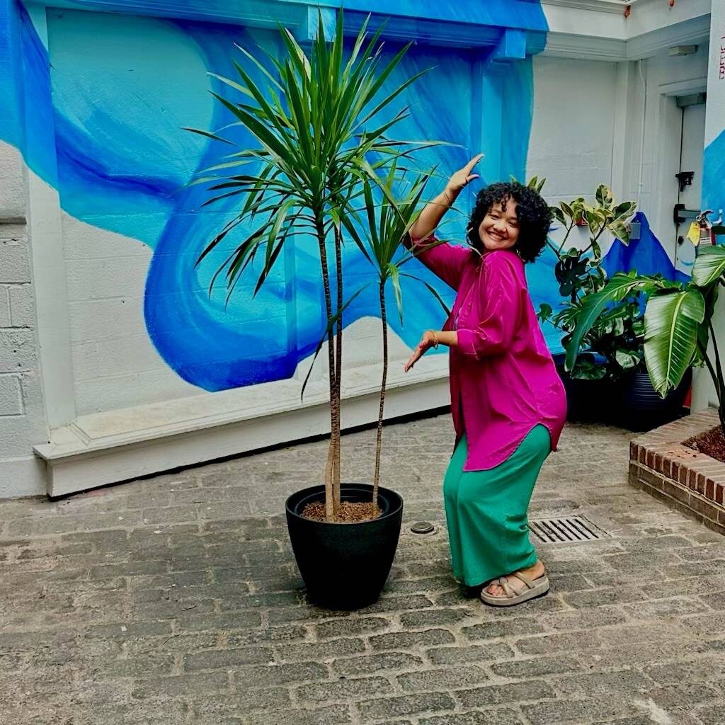 Visitor posing with a potted dragon tree beside the mural at Stem & Vine in Baltimore